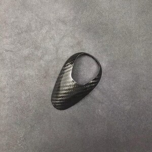 May include: A black carbon fibre gear shift knob with a loop handle. The knob has a woven pattern and is set against a grey background. The shape is ergonomic and designed for easy gripping.