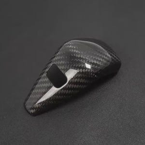 May include: A black carbon fibre car gear shift knob. The knob has a glossy finish and a woven pattern. It features a rectangular opening on the front. The background is a dark grey colour.