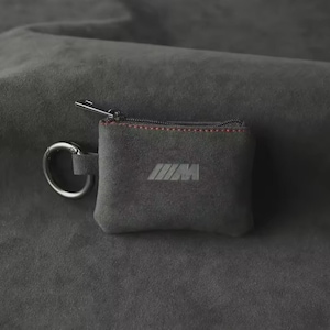 May include: A small, grey suede key pouch with a silver zip and a silver key ring. The pouch features the embossed letters "M" and "W" in a stylized design. The zip is accented with red stitching.