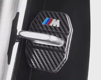 May include: A carbon fiber car door lock cover with the BMW M logo in blue, red, and white. The cover has a textured, woven pattern and a silver-colored latch.
