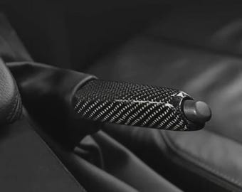 May include: A close-up of a black car handbrake with a carbon fiber weave pattern. The handbrake is in a car interior, with black leather seats visible in the background. The handbrake has a black button on the end.