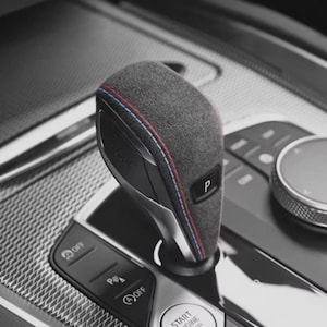 May include: A close-up of a car's gear shift lever. The lever is grey with red and blue stitching. The letter "P" is visible on the lever. The surrounding console is black and silver.