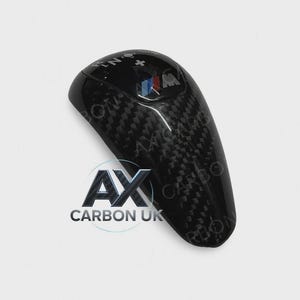 May include: A black carbon fibre gear shift knob with a glossy finish. The knob has a textured pattern and features the BMW logo. The text "N" and "+" are visible on the top. The logo "AX CARBON UK" is at the bottom.