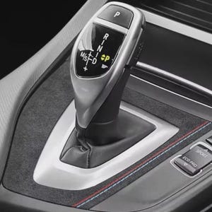 May include: A silver car gear shift lever with black markings for Park, Reverse, Neutral, Drive, and Manual/Sport modes. The surrounding console is black with a textured finish and features red, white, and blue stitching as a design element.