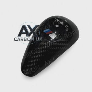 May include: A black carbon fibre gear shift knob with the BMW logo in red, blue, and white. The knob has gear selection indicators and the text "AX CARBON UK".