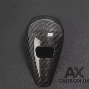 May include: A black carbon fibre car key fob cover with a glossy finish. The cover features a rectangular opening. The text "AX CARBON UK" is visible in white. The key fob cover has a woven pattern.
