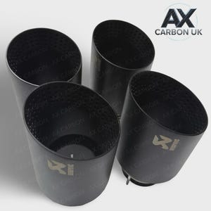 May include: Four black exhaust tips with angled openings. The tips have a perforated design near the opening and feature the text "AX CARBON" and a logo. The background is a light grey.