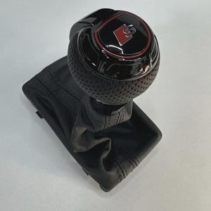 May include: A black gear stick knob with a perforated grip and a red and black emblem. The gear stick is attached to a black leather boot with detailed stitching. Automotive part.
