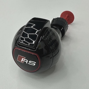 May include: Black and carbon fibre car gear shift knob with a red and white RS logo. The knob has a geometric design on top and a red circular piece at the end.