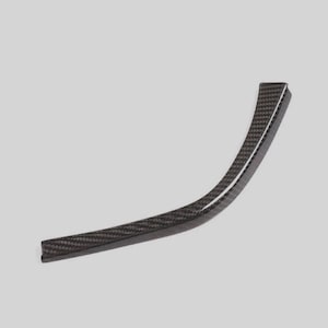 May include: A curved, black carbon fibre trim piece with a woven pattern. The trim has a glossy finish and a channel along one edge. The shape is a gentle arc, suitable for automotive or decorative applications.