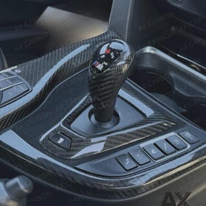 May include: Carbon fibre gear shift knob and console trim. The gear shift knob has a glossy finish with the M logo. The console features various buttons and a textured carbon fibre pattern. The text "AX CARBON" and "CARBON UK" are visible.