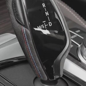 May include: A close-up of a black and grey car gear shift lever. The lever has a textured grip with red and blue stitching. Gear positions are labelled P, R, N, D, and M/S. The base of the lever is surrounded by a glossy black surface.