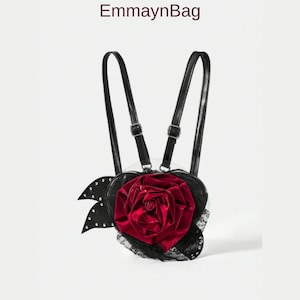 Puede incluir: Mochila negra en forma de corazón con una gran rosa roja en el centro. La mochila tiene correas negras y un ribete de encaje negro. El texto "EmmaynBag" está en la parte superior.