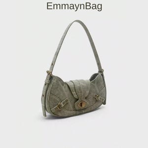 May include: A sage green shoulder bag with a curved shape and a flap closure. The bag features a long strap, buckle details, and a gold-toned clasp. The text "EmmaynBag" is visible at the top.