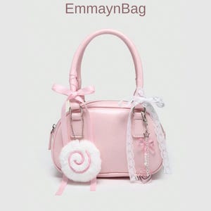 May include: A pale pink handbag with a rounded rectangular design, featuring a top handle and a small zip. The bag is decorated with pink ribbons, a white fluffy charm, and a decorative keychain with a star and beads. The text "EmmaynBag" is at the top.