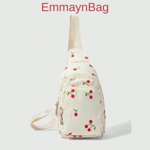 Op de afbeelding: Crèmekleurige corduroy sling bag met een kersenpatroon. De tas heeft een klein voorvak, een ritssluiting en een crèmekleurige band. De tekst "EmmaynBag" staat in het rood bovenaan.