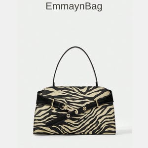 Puede incluir: Bolso de mano rectangular con estampado de cebra en negro y beige. Tiene un asa negra y una correa negra con herrajes dorados. El texto "EmmaynBag" es visible en la parte superior de la imagen.