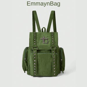 Può includere: Zaino verde con chiusura a patta, tasche laterali e spallacci regolabili. Lo zaino presenta un emblema a croce decorativo e borchie metalliche. Il marchio "EmmaynBag" è stampato in alto.