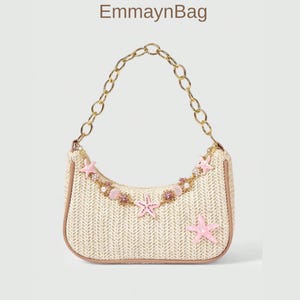 Puede incluir: Un bolso beige tejido con una correa de cadena dorada. El bolso está decorado con estrellas de mar rosas y piedras preciosas. El texto "EmmaynBag" está en la parte superior. El bolso tiene forma curva y ribete marrón.