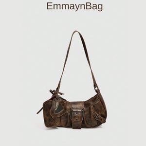 Puede incluir: Bolso de hombro de ante marrón con una correa larga. El bolso tiene un cierre de solapa con hebilla metálica y un pequeño bolsillo delantero. Una pequeña etiqueta con ventana transparente está adjunta. El texto "EmmaynBag" es visible.