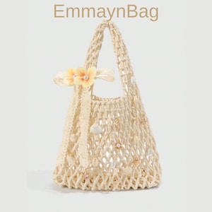 May include: Cream-colored, open-weave tote bag with a floral embellishment and seashell accents. The bag features a handle and is adorned with a cream-colored lace ribbon. The text "EmmaynBag" is at the top.