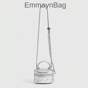 Puede incluir: Un bolso bandolera plateado acolchado con asa superior y correa de cadena. El bolso tiene forma pequeña y redonda y cierre de cremallera. El texto "EmmaynBag" está en la parte superior de la imagen.