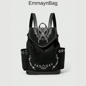 Puede incluir: Una mochila negra con el texto EmmaynBag en la parte superior. La bolsa presenta un diseño estampado en blanco de una polilla, fases lunares, estrellas y ramas con hojas en la solapa y el cuerpo. Incluye dos bolsillos laterales con ojales metálicos plateados.