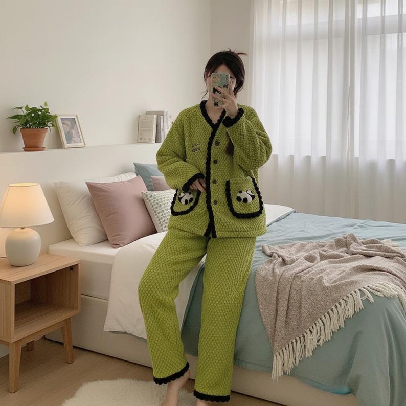 May include: Green pajama set with a textured pattern. The jacket has black trim, buttons, and panda bear pocket details. The trousers have black trim at the ankles. The pajamas are made of a soft, plush material.