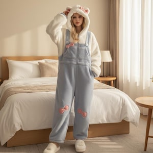 May include: A cozy, light blue and white jumpsuit with a hood featuring bear ears. The jumpsuit has pink bow accents and a zip-up front. The outfit is made of a soft, plush material, perfect for lounging or sleepwear.