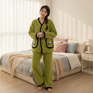 May include: A green pajama set with a textured pattern. The jacket has black trim, panda bear pocket details, and the text "Sweet Dreams". The set includes a button-up jacket and matching trousers. The person is holding a white cup.