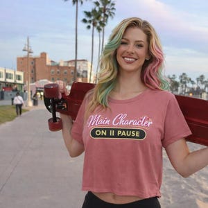 Puede incluir: Una mujer con un top corto rosa con el texto "Main Character ON II PAUSE" sostiene una patineta roja. Su cabello rubio tiene mechones rosas, verdes y azules. Lleva leggings negros y sonríe. Hay palmeras y edificios al fondo.