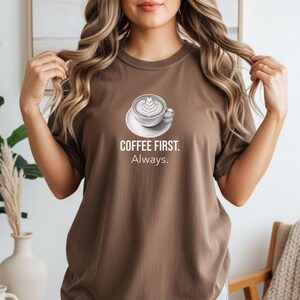 May include: A brown t-shirt featuring the text "COFFEE FIRST. Always." and a coffee cup graphic with latte art. The shirt is plain and has short sleeves.