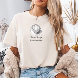 May include: Cream-colored t-shirt featuring a grayscale coffee cup design with latte art. The text "Same Shop Same Order" is printed below the graphic. The shirt is made of a soft material.