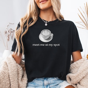 May include: Black t-shirt featuring a grayscale image of a coffee cup with latte art on a saucer. The text "meet me at my spot" is printed below the coffee cup. The shirt is a casual style.