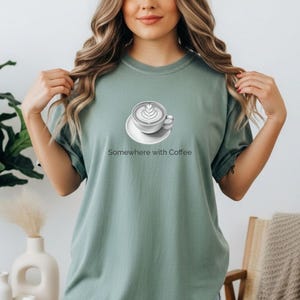 May include: A white ceramic mug with a black handle and the text "But First, Coffee" in a handwritten style. The mug is on a white surface. The mug is approximately 10 cm tall.