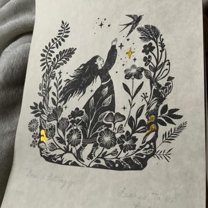 May include: Black and white linocut print of a woman reaching for a bird, surrounded by flowers and foliage. The print has gold foil accents and the text "Love is Letting go". Signed by the artist.
