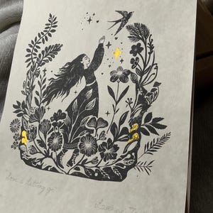 May include: Black and white art print featuring a woman reaching for a bird, surrounded by floral elements. Gold accents highlight stars and details. The text "Love is letting go" is visible, along with the artist's signature.