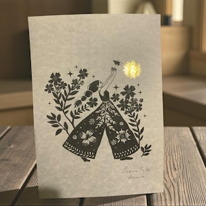 May include: A greeting card featuring a black and white illustration of a woman reaching for a butterfly and a sun. The woman is surrounded by flowers and foliage. The card has the text "Leanne To the Anamaria" at the bottom.