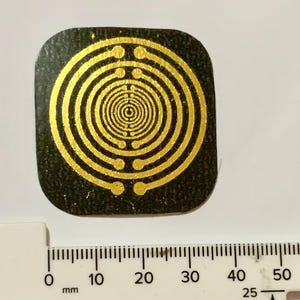 Pode incluir: Um objeto quadrado, de cor escura, com um design de círculos concêntricos dourados. O design é centrado e detalhado, com uma régua mostrando milímetros. O objeto tem aproximadamente 4 cm de largura.