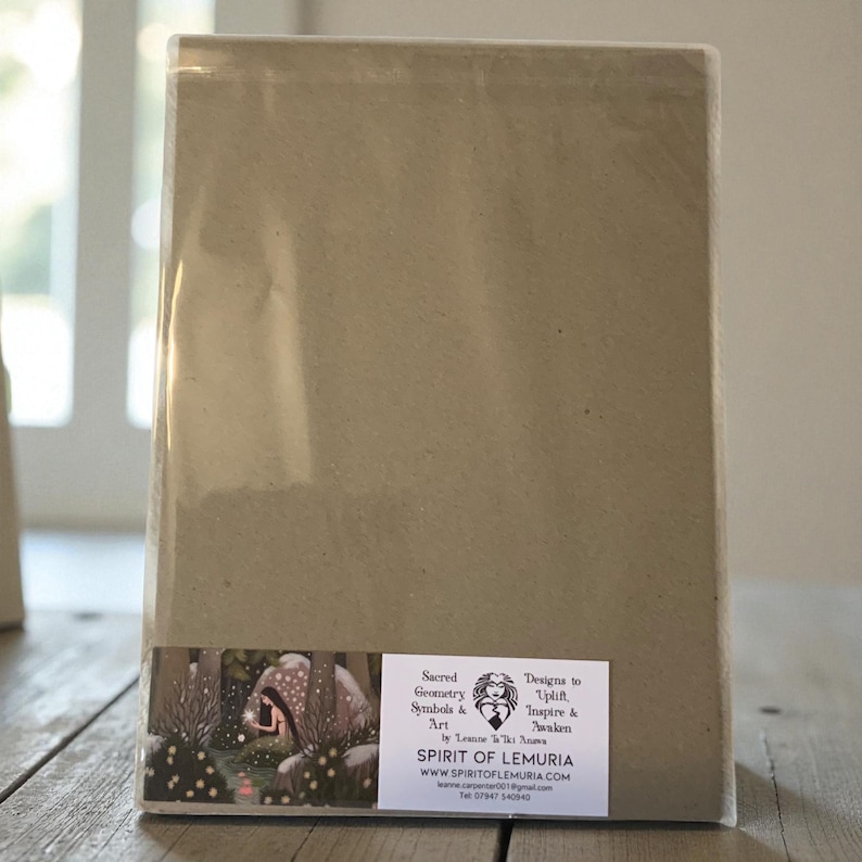 May include: A rectangular package wrapped in clear plastic. The package has a light brown paper backing with a sticker featuring an illustration of a forest scene and text that reads "Sacred Geometry Symbols & Art" and "Spirit of Lemuria."