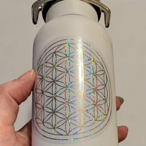 May include: White stainless steel water bottle with a silver hinged lid and a holographic flower of life design. The design has a rainbow sheen. The bottle is approximately 8 inches tall.