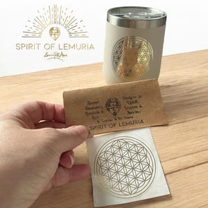 Può includere: Un bicchiere color crema con un design geometrico dorato e un coperchio argentato. Sono visibili anche una piccola carta con la scritta "Spirit of Lemuria" e un adesivo abbinato. Il logo presenta un design a raggiera e le parole "Spirit of Lemuria".