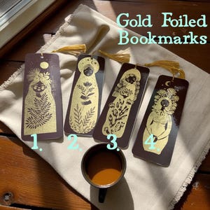 Può includere: Quattro segnalibri dorati con dettagli a nappa. Ogni segnalibro presenta un'illustrazione unica di una figura con dettagli dorati su sfondo marrone scuro. Il testo "Gold Foiled Bookmarks" è visibile.