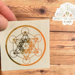 May include: A round sticker with a gold and orange Metatron's Cube design. The sticker is held by a hand and has a white background. The sticker also has the text "SPIRIT OF LEMURIA" in a stylized font.