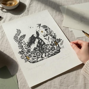 May include: Black and white art print featuring a woman reaching for a bird, surrounded by floral elements. The print includes the text "Love is a letting go." and is held near a cup of tea and glasses.