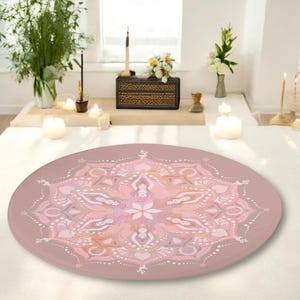 Pode incluir: Um almofada de meditação redonda rosa com um design de mandala em tons de rosa, branco e dourado. O almofada está sobre uma superfície branca.