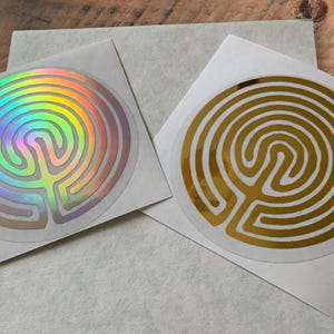 Pode incluir: Dois autocolantes circulares com um desenho de labirinto. Um autocolante tem um acabamento holográfico, colorido com o arco-íris, enquanto o outro é dourado. Ambos estão em papel branco, com um fundo neutro.
