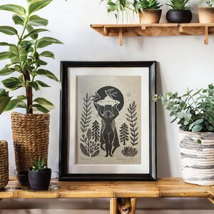 May include: A framed black and white art print featuring a stylized figure holding a celestial object, surrounded by botanical elements. The print is displayed on a bamboo shelf, alongside potted plants in woven baskets and ceramic pots, creating a natural aesthetic.