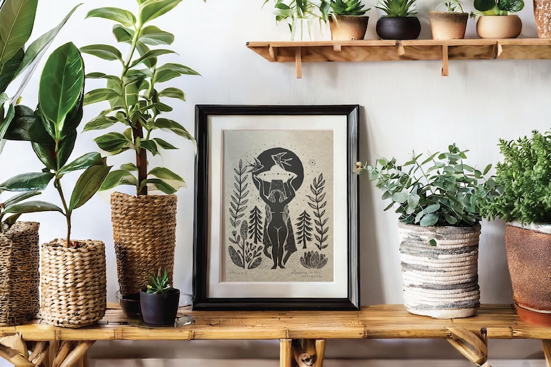 May include: A framed black and white art print featuring a stylized figure holding a celestial object, surrounded by botanical elements. The print is displayed on a bamboo shelf, alongside potted plants in woven baskets and ceramic pots, creating a natural aesthetic.