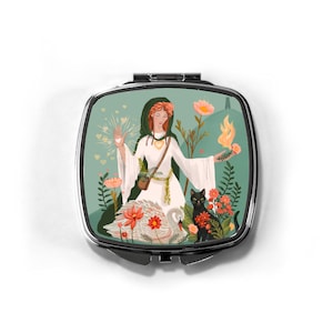 May include: A compact mirror with a silver frame and a colorful illustration of a woman in a white dress and green cloak, surrounded by flowers, a swan, and a black cat. The woman is holding a heart and a flame.